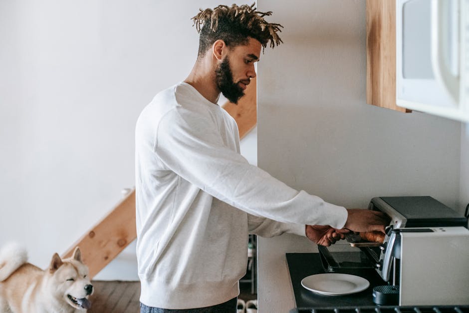 Side view of stylish young African American male owner in casual clothes heating croissant in microwave oven while standing in kitchen with cute Akita Inu dog