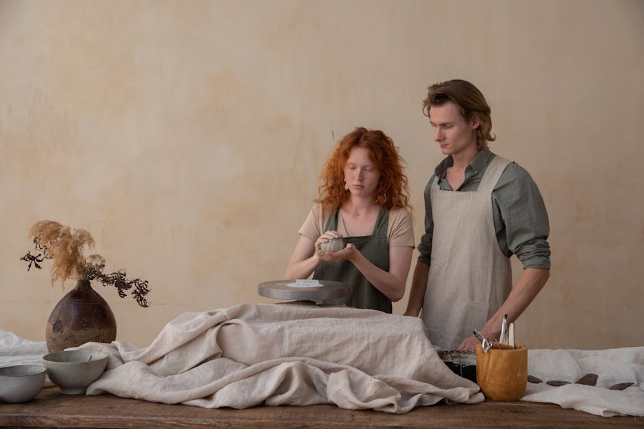 Focused couple in aprons modelling clay plates together while standing near wooden table with supplies
